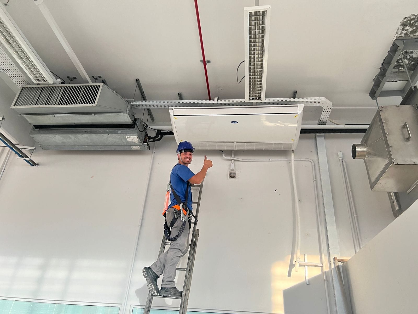 técnico com capacete azul e cinto de segurança em cima de uma escada dando joinha ao lado de um ar-condicionado cassete 