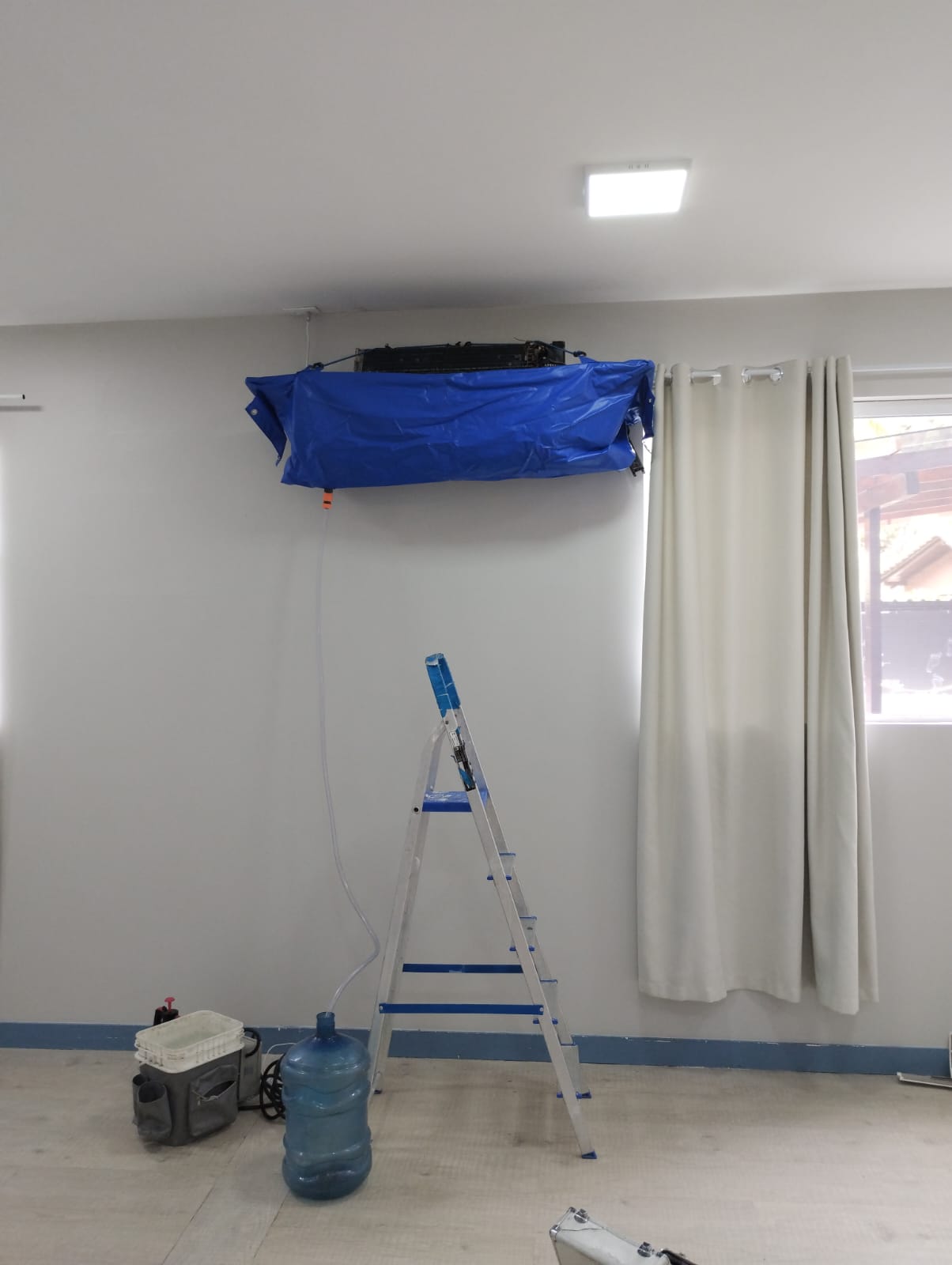 ar-condicionado split na parede coberto por lona azul durante limpeza, com mangueira drenando para um galão de